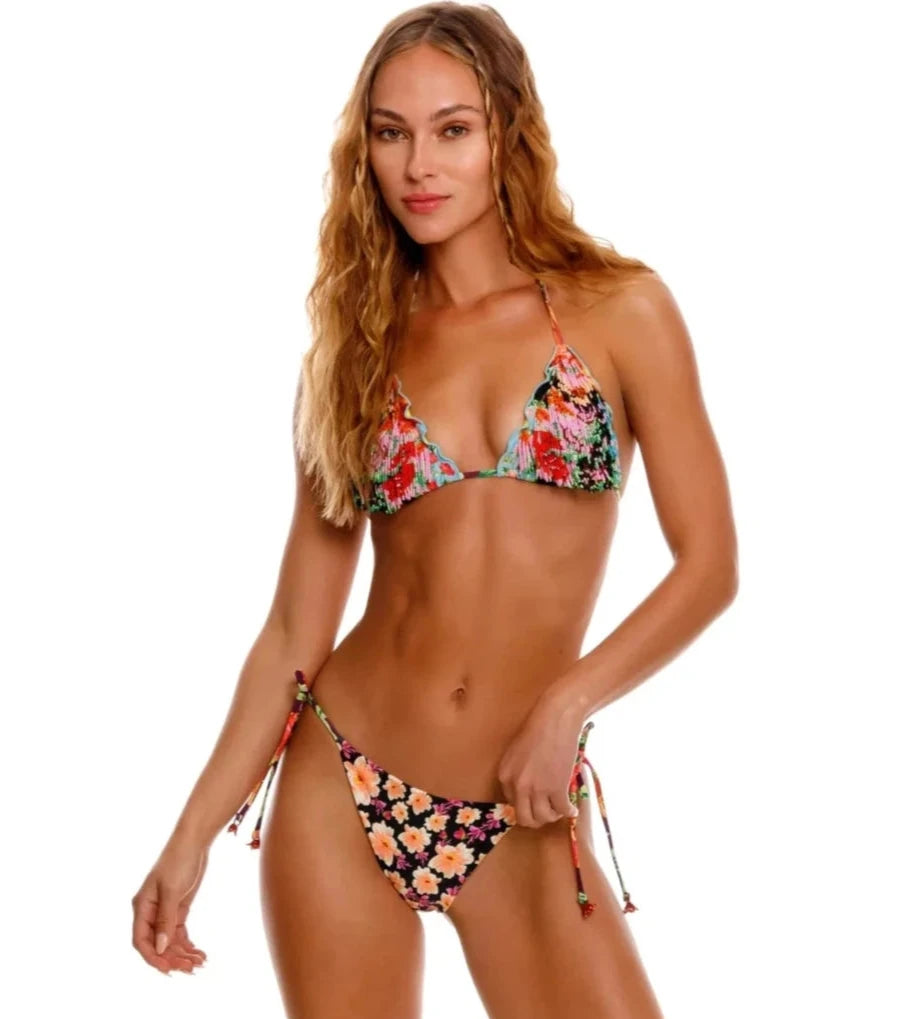 A woman wearing a floral triangle bikini top with scalloped edges and matching string-tie bottoms, posing with relaxed confidence.