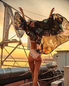 Woman facing sunset wearing peach colored high waisted bikini bottoms and black sarong in the air behind her.